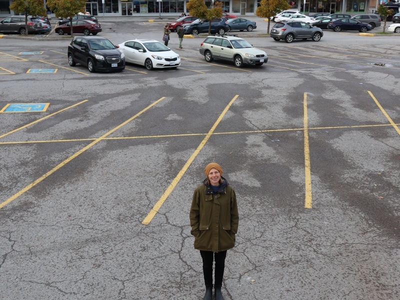 A woman with her hands in her pockets stands in the middle of a wide open parking lots, with some cars parked in the background.