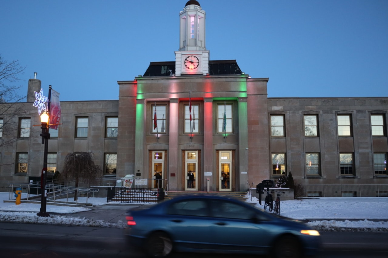 Photo shows Peterborough city hall lit up in the evening, with a car passing on the street in front.