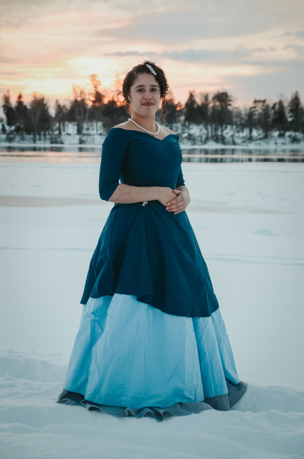 Photo shows woman in a long dress standing in front of the frozen Otonabee River.
