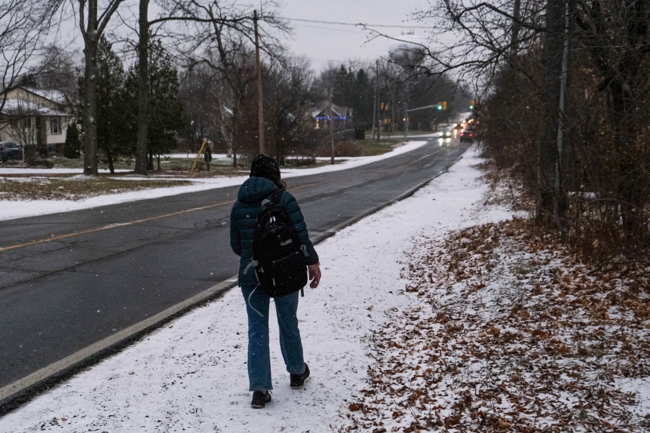 Photo shows a woman walking on the gravel shoulder of Sherbrooke Street West, on a snowy day.