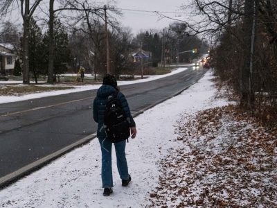 Photo shows a woman walking on the gravel shoulder of Sherbrooke Street West, on a snowy day.
