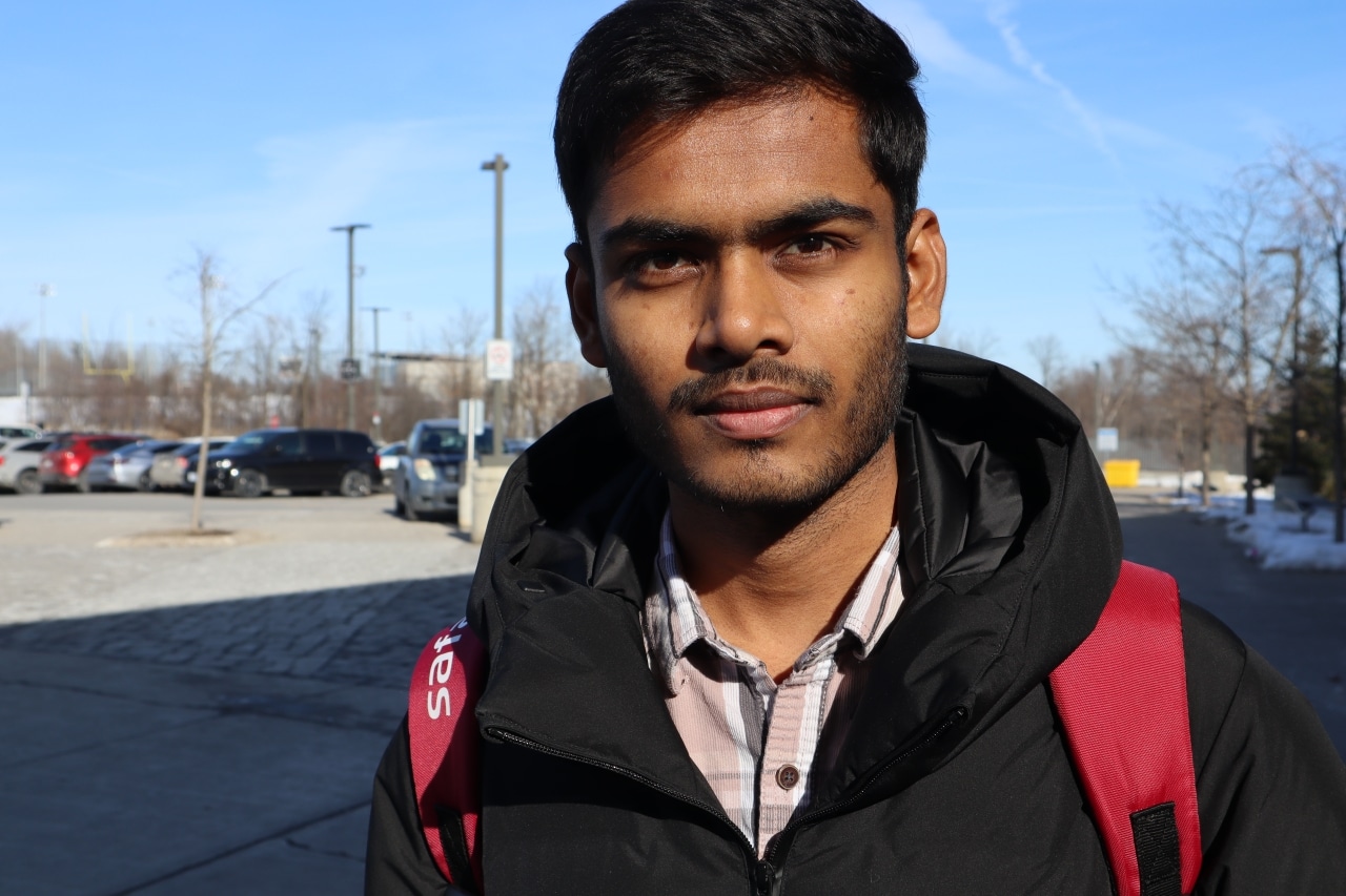 Photo shows a young man with short hair wearing a winter coat and a backpack while standing in front of a parking lot.