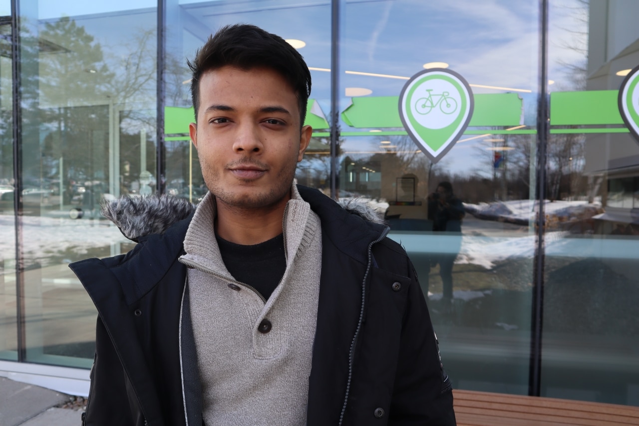 Image shows a young man with short hair standing outdoors in front of a bank of large windows.