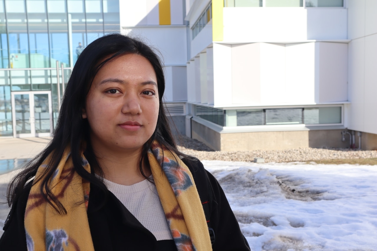 Photo shows a young woman with shoulder-length hair standing in front of a building at Fleming College's Peterborough campus.