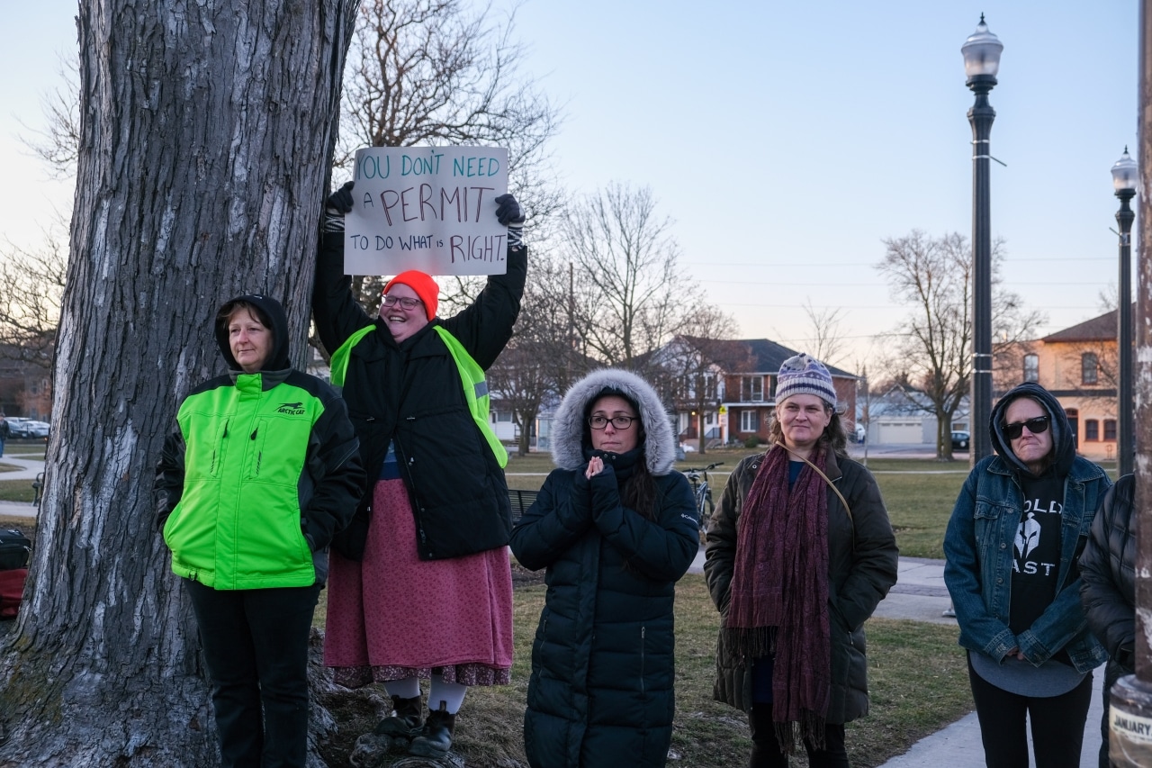 Several people stand in Confederation Square, while one of them smiles and holds high a sign that reads "you don't need a permit to do what is right." 