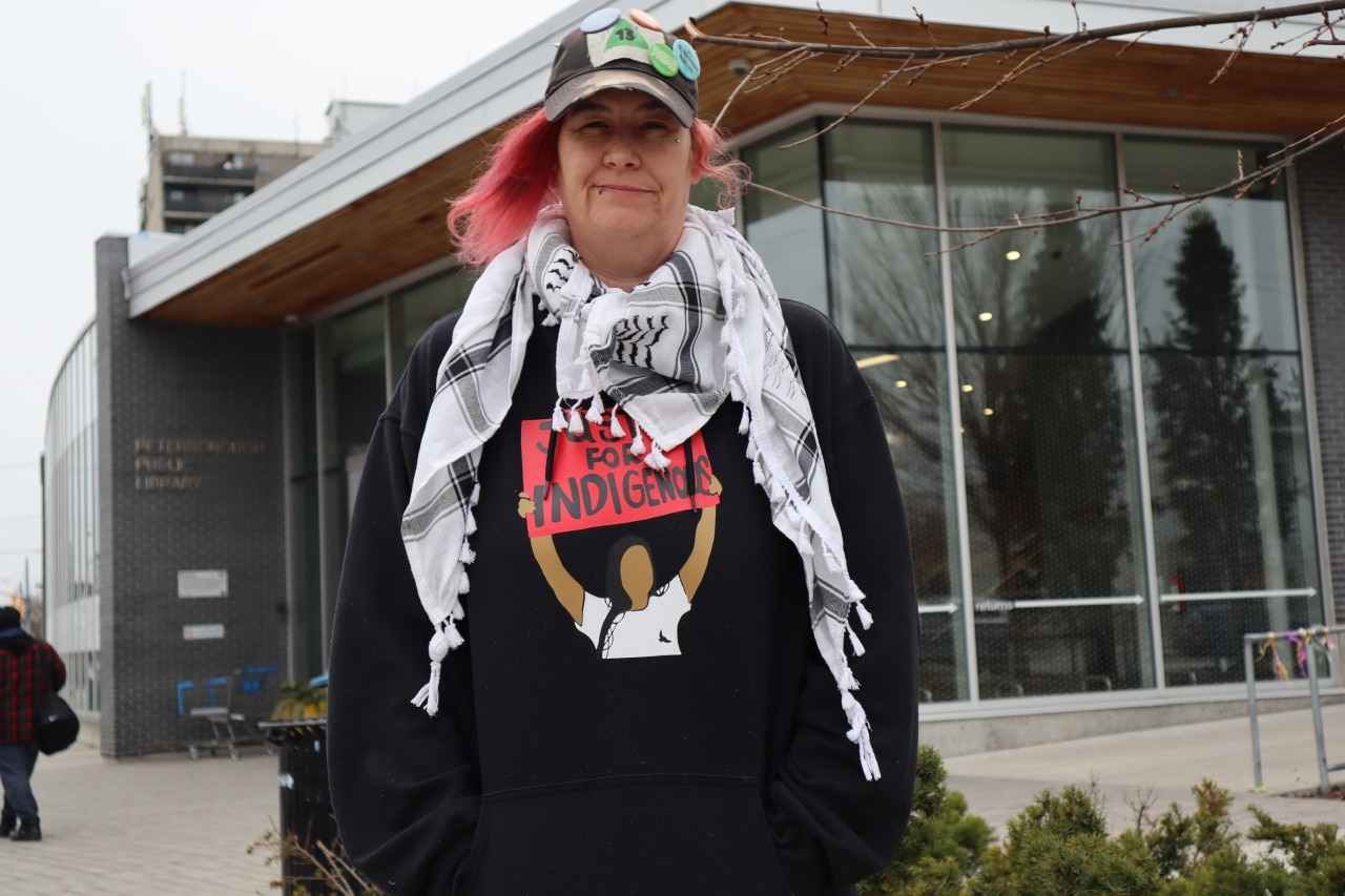 Photo shows Sam Blondeau wearing a ball cap over pink hair and a keffiyeh over a black sweatshirt that says "Justice for Indigenous," while standing in front of the entrance of the Peterborough Public Library.