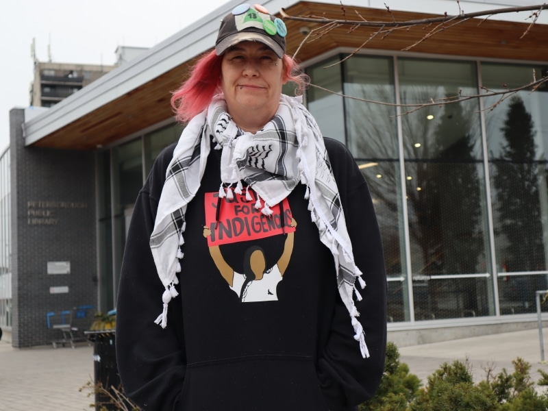 Photo shows Sam Blondeau wearing a ball cap over pink hair and a keffiyeh over a black sweatshirt that says "Justice for Indigenous," while standing in front of the entrance of the Peterborough Public Library.