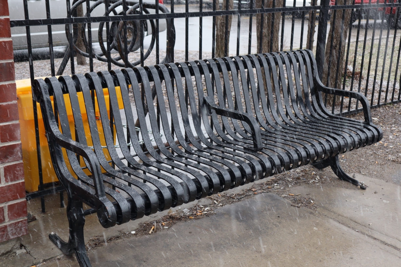 Photo shows snow falling on a black metal bench that has an armrest on either end and another in the middle.