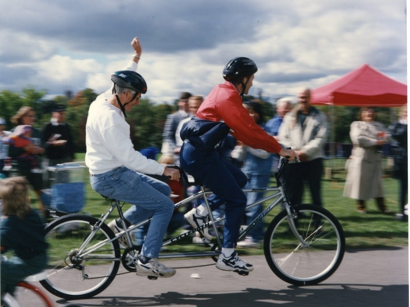 Photo shows two people riding a tandem bicycle on the Rotary Trail past a group of people gathered in Rotary Park. The person on the back is raising their fist in the air.