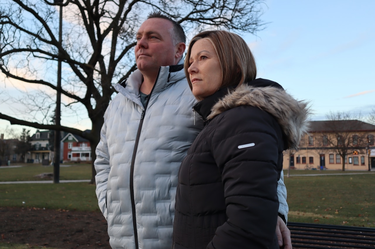 Photo shows a couple in winter coats standing in a park in the late afternoon on a December day.