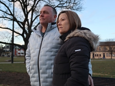 Photo shows a couple in winter coats standing in a park in the late afternoon on a December day.